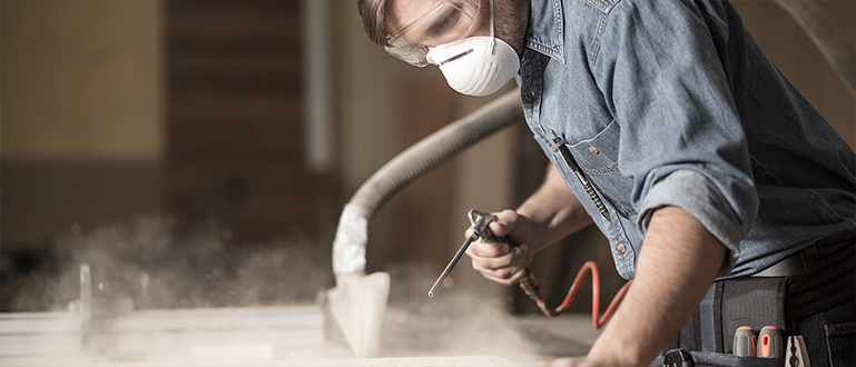 Tischler mit Mund-Nasen-Schutz entfernt Holzstaub von einem Brett Tischler mit Mund-Nasen-Schutz entfernt Holzstaub von einem Brett